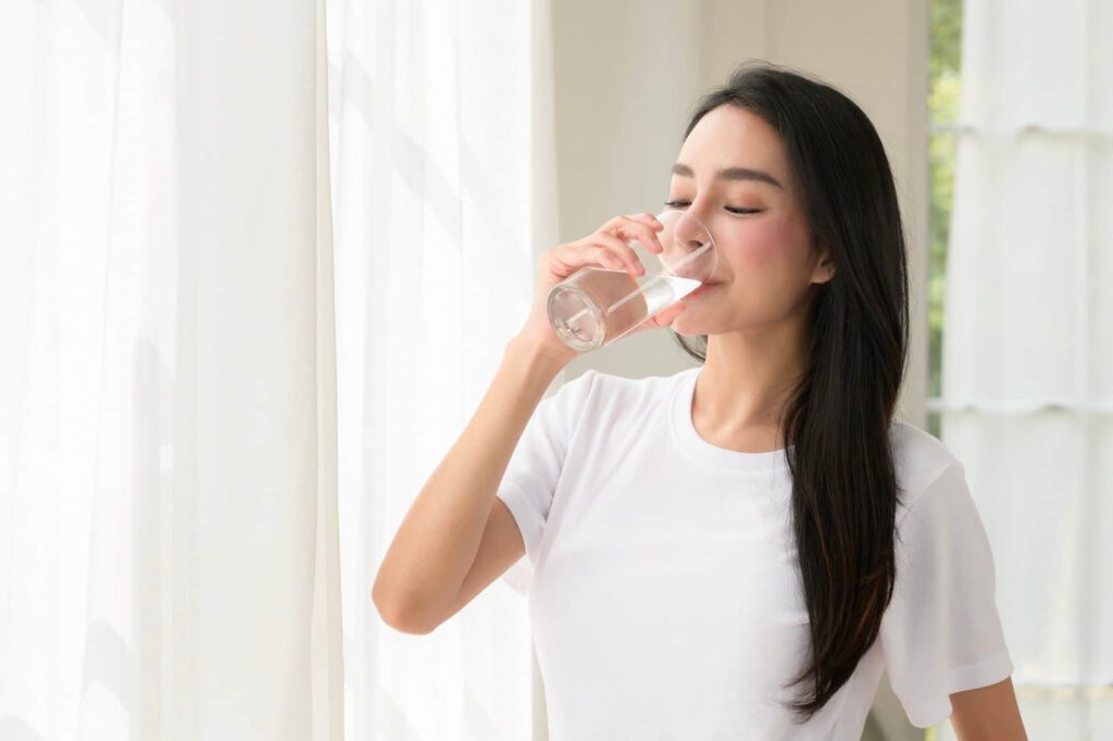 Woman drinking water indoors by a sunny window.
