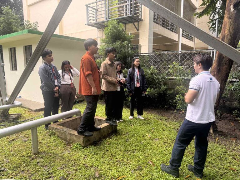 A group of students listens to an instructor in a grassy outdoor area.